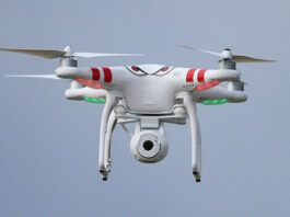 White coloured drone with red stripes in the air against a pale blue sky.