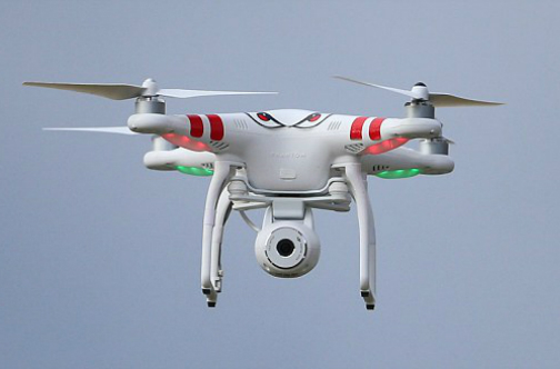 White coloured drone with red stripes in the air against a pale blue sky.