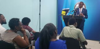 Prime Minister Philip J. Pierre stands at a podium fielding questions from seated journalists.