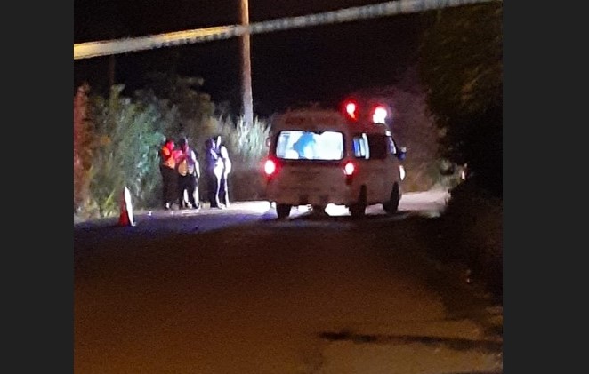 Police caution tape hovers above an ambulance in the darkness at the scene of a homicide in Babonneau, while curious onlookers stand at the side of the road.