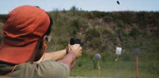 Man wearing cap and ear plugs fires handgun at target in outdoor shooting range.