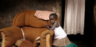 Minor female leans on a broken-down chair in a poor household.