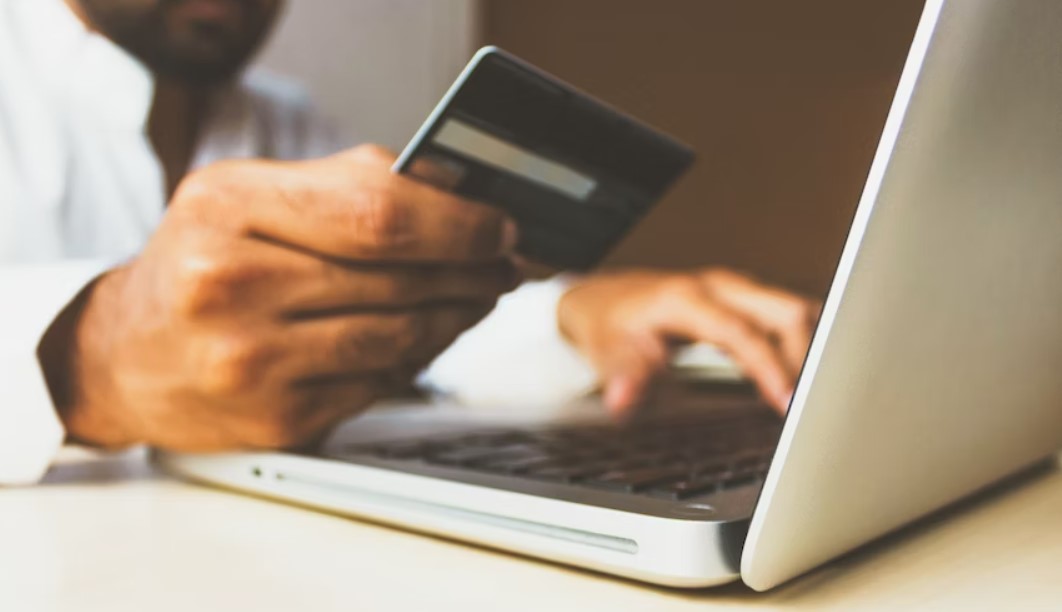 Man holds credit card in one hand while typing on computer with the other.