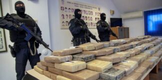 Masked officers dressed in black with assault rifles stand guard over multiple packages of illegal drugs.