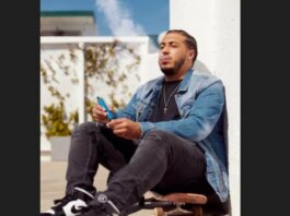 Man sits in a corner near a skateboard smoking an e-cigarette.