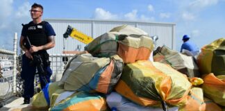 Coast Guard official with automatic weapon stands guard over bales of seized cocaine.