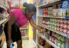 Female consumer examines canned goods on supermarket shelf.