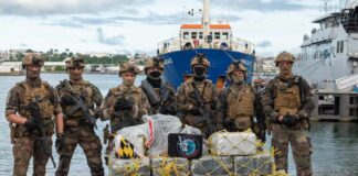 Armed French law enforcement officers stand against the backdrop of navy vessels with bales of seized cocaine on the ground in front of them.