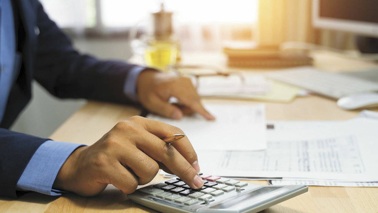 Man in blue suit sits at a table with several sheets of paper doing calculations on an electronic calculator.