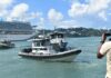Television cameraman points camera at Marine Police vessel at sea with cruise ship in the background.