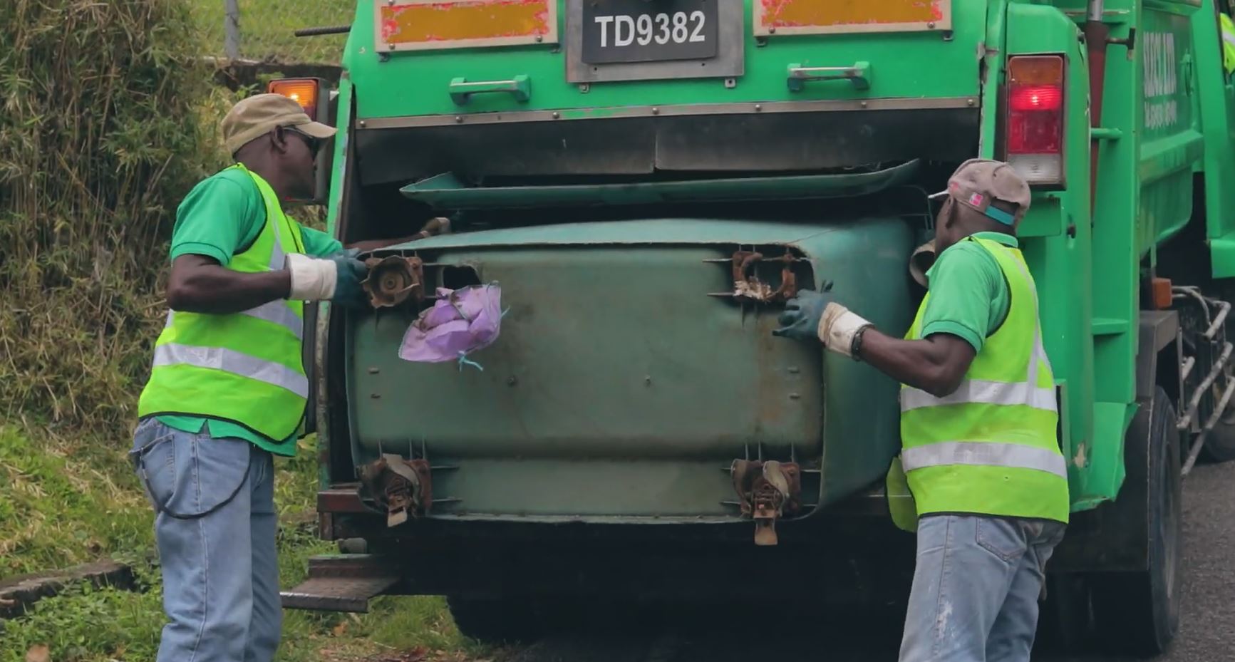 Two garbage collectors dressed in green reflective clothing empty garbage into a garbage truck.