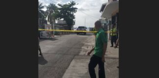 Man stands in front of police caution tape behind which armed officer stands guard at homicide scene in Gros Islet.
