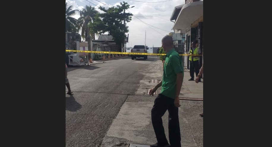 Man stands in front of police caution tape behind which armed officer stands guard at homicide scene in Gros Islet.
