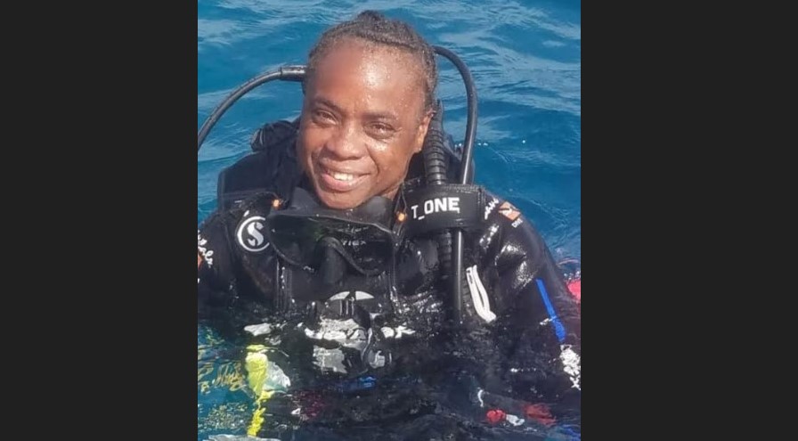 Vanessa Eugene dressed in diving wetsuit with breathing apparatus on her back emerges smiling from the water.