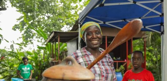 Woman with wooden spoon in hand in front of earthenware vessel.