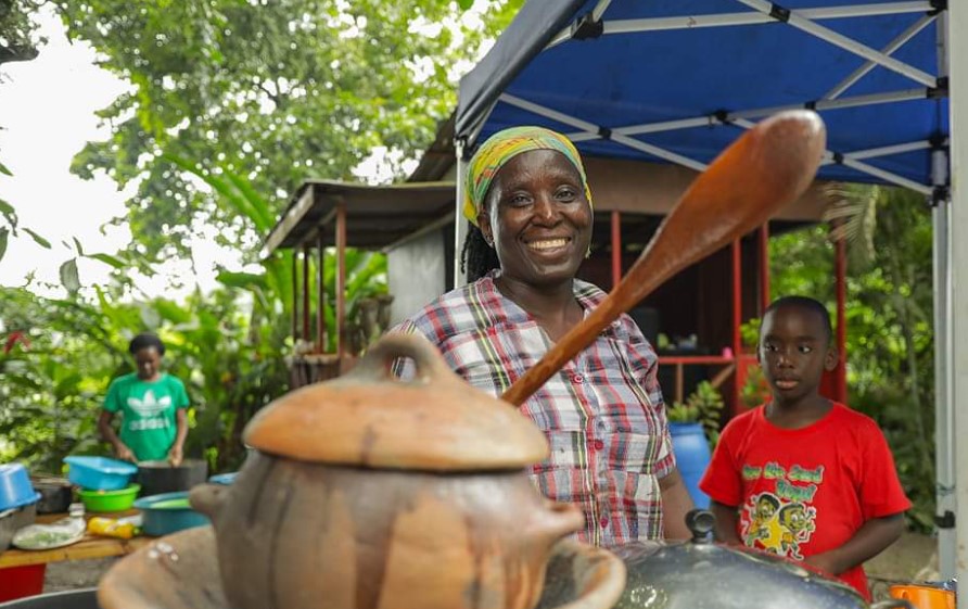 Woman with wooden spoon in hand in front of earthenware vessel.